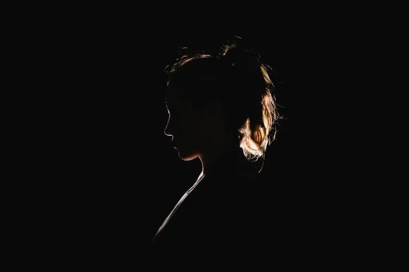 A woman’s silhouette is shown in profile against a black background, with light illuminating the edges of her hair and face, creating a dramatic, high-contrast effect.