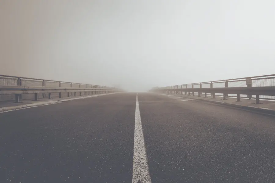 A straight, empty road with metal guardrails on both sides disappears into thick fog, creating a sense of mystery and calm. The scene is devoid of vehicles or people.