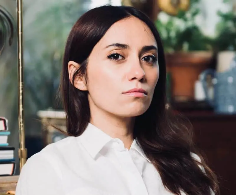 A woman with long dark hair and a serious expression is wearing a white shirt. She is sitting indoors with blurred plants and books in the background.