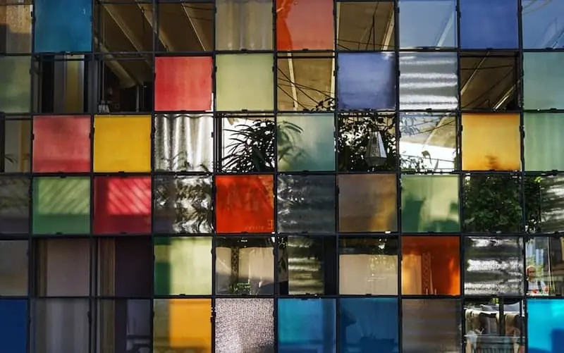 A grid of colorful glass panels, including shades of red, yellow, blue, and green, forms a wall. Plants and reflections are visible behind the transparent and translucent sections.