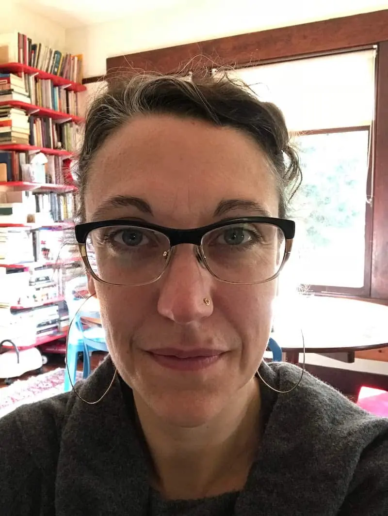 A person with glasses and hoop earrings is looking at the camera. Behind them are shelves filled with books, a table, chairs, and a large window letting in natural light.