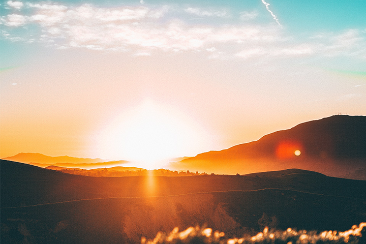 Bright orange sunrise over rolling hills and mountains, with sun rays illuminating the landscape and a partly cloudy sky above. The scenery appears serene and the atmosphere glows warmly.