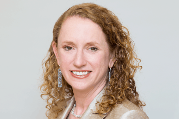 A woman with curly, light brown hair smiles at the camera. She is wearing a light-colored blazer, dangling earrings, and a necklace, with a neutral background behind her.