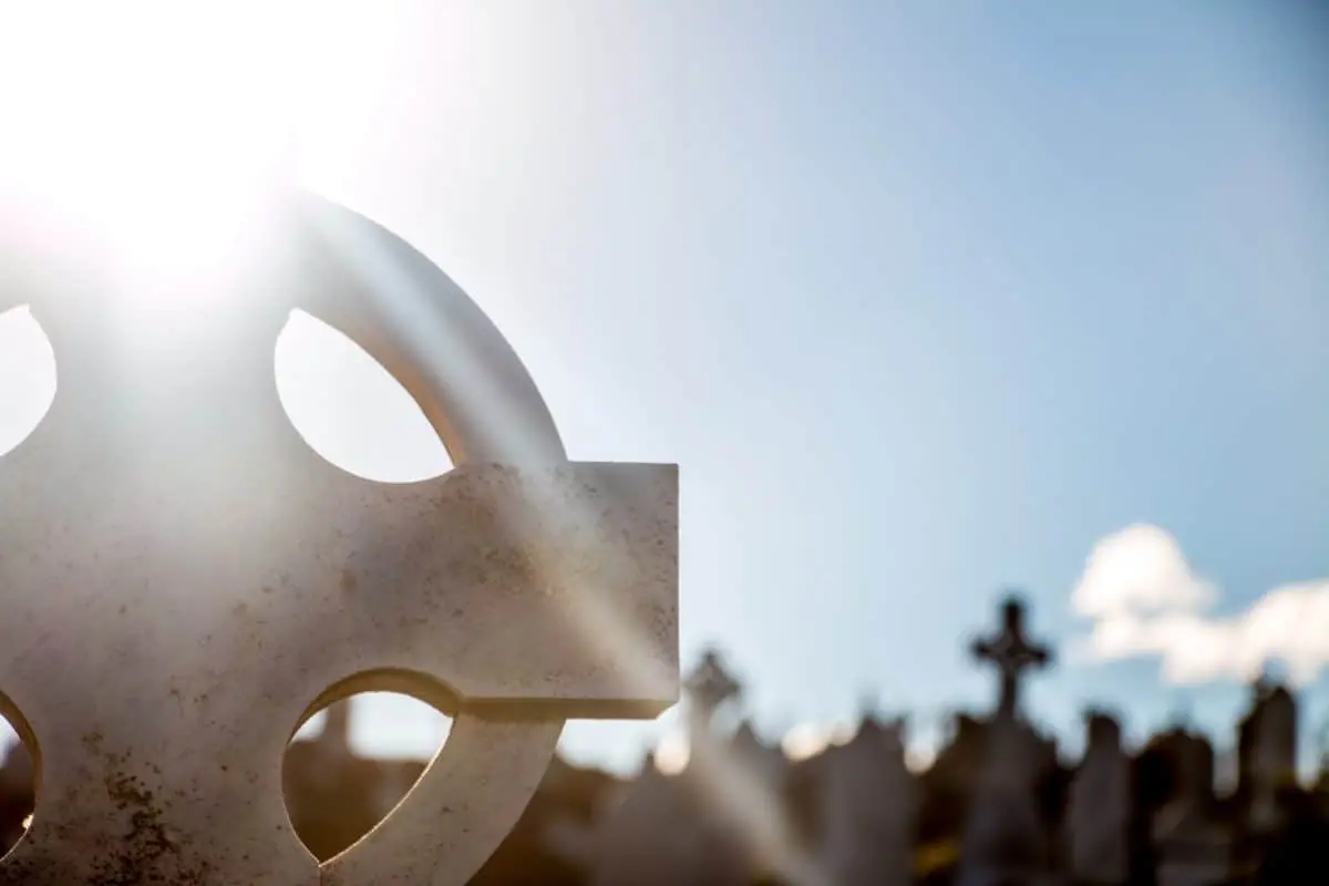 A close-up of a Celtic cross headstone in a cemetery, with sunlight shining brightly from the left, creating lens flare and silhouetting other crosses against a blue sky in the background.