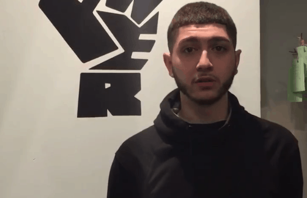 A young man with short dark hair and a trimmed beard stands in front of a wall with a bold black graphic of a raised fist and block letters. He is wearing a black hoodie and looking slightly past the camera.