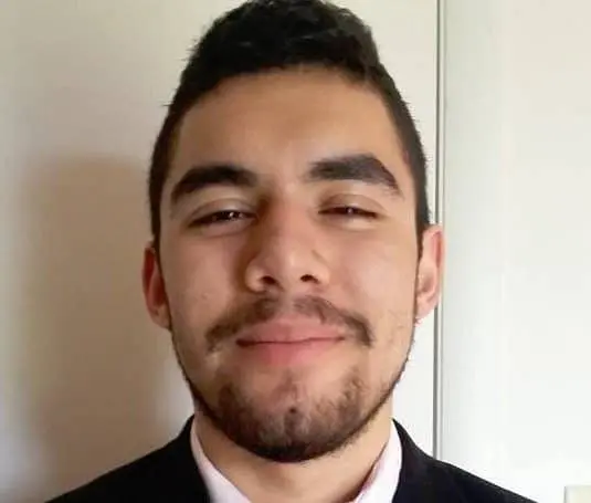 A young man with short dark hair, a trimmed beard, and mustache is smiling slightly. He is wearing a black suit jacket and standing in front of a light-colored wall.