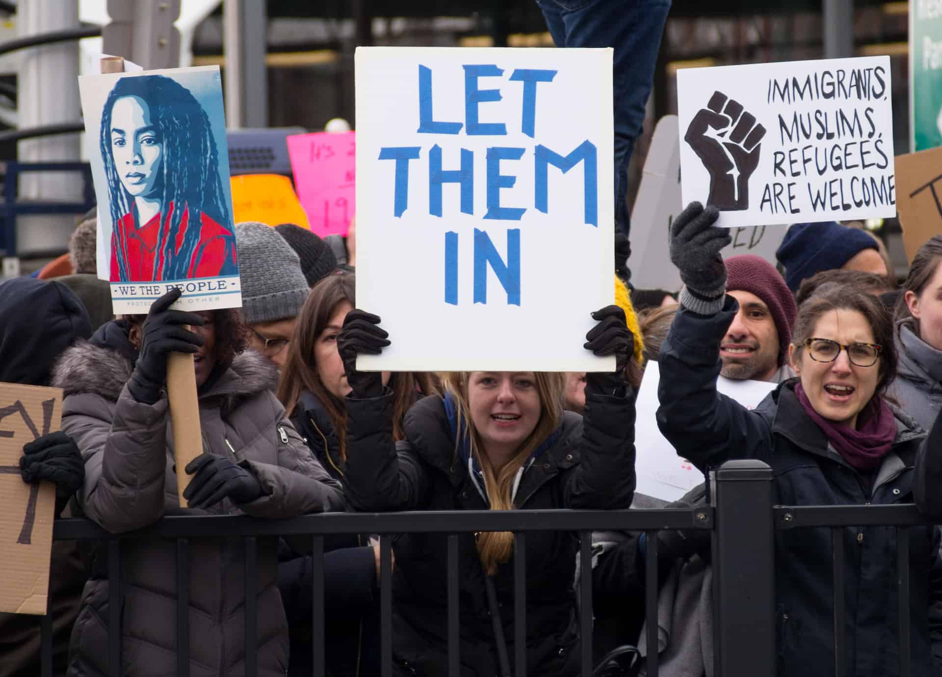 A group of protesters stand outside holding signs, including one that says LET THEM IN and another that reads IMMIGRANTS MUSLIMS REFUGEES ARE WELCOME with a raised fist symbol.