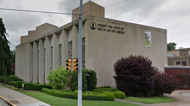 A beige building with large windows and the sign Tree of Life – Or L’Simcha in English and Hebrew on the wall, surrounded by green bushes and a manicured lawn, under a cloudy sky.
