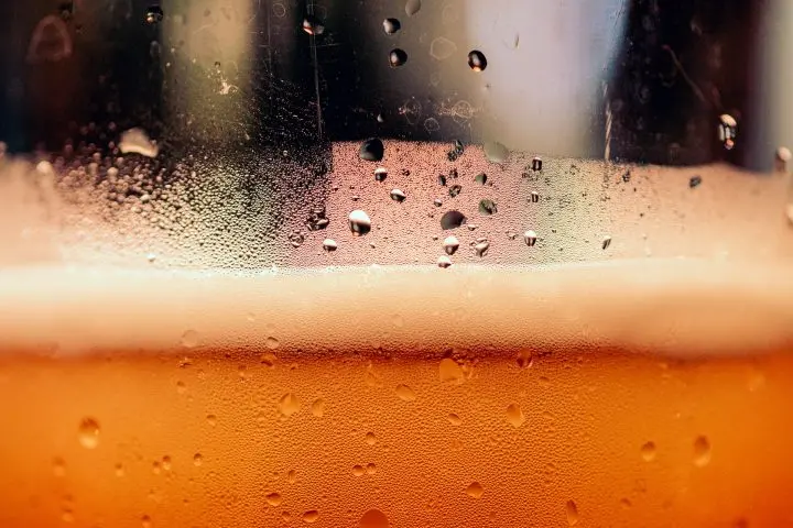Close-up of a glass of beer with condensation and droplets on the outside, a layer of foam at the top, and an amber liquid below.