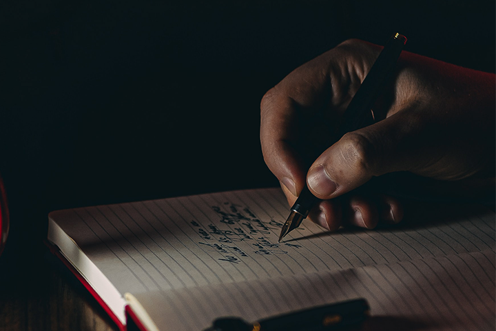 A close-up of a hand holding a pen and writing notes in a lined notebook, with dim lighting creating a focused and intimate atmosphere.