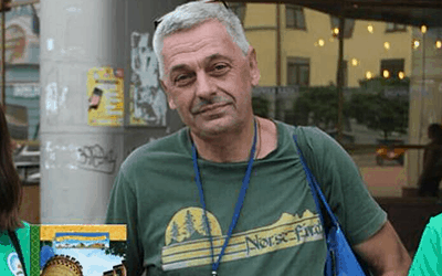 A middle-aged man with short gray hair wears a green Norse-Find T-shirt and a blue lanyard, standing outdoors in front of a building with large windows. He carries a blue bag over his shoulder and looks at the camera.