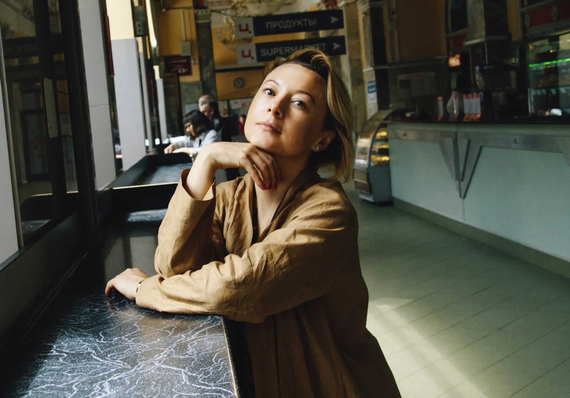 A woman in a tan coat sits at a marble counter by a window in a brightly lit cafe, her chin resting on her hand as she gazes thoughtfully at the camera—reminiscent of a candid moment from a Valzhyna Mort interview. Signs and counters fill the background.