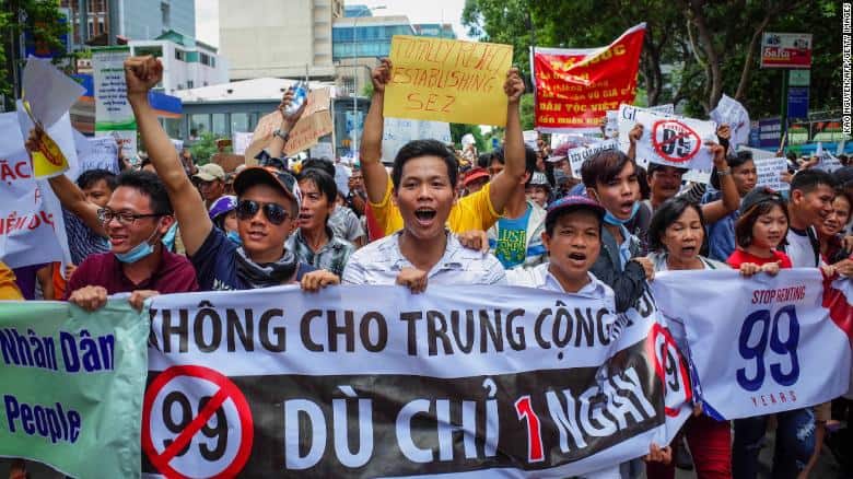 A crowd of protesters march holding signs and banners, including one that says KHÔNG CHO TRUNG CỘNG THUÊ 99 NĂM, DÙ CHỈ 1 NGÀY! with symbols opposing a 99-year lease agreement. The atmosphere is energetic and passionate.