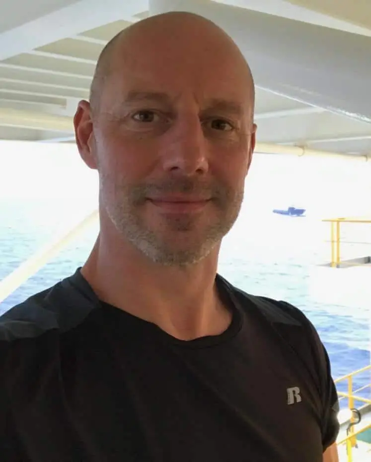A bald man wearing a black T-shirt is smiling at the camera on a boat, with the ocean and a distant boat in the background—capturing the adventurous spirit of Will Mackin’s Pen Ten stories.