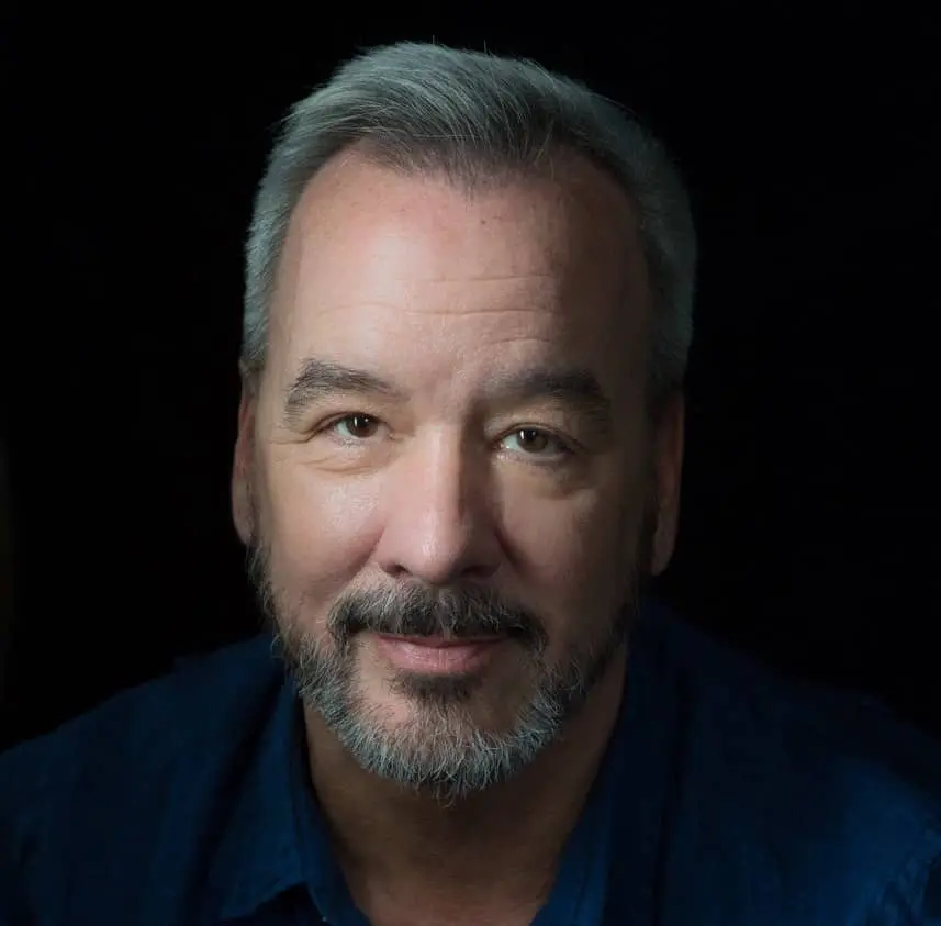 A middle-aged man with short gray hair and a beard, wearing a dark blue shirt, smiles softly while looking directly at the camera against a dark background.
