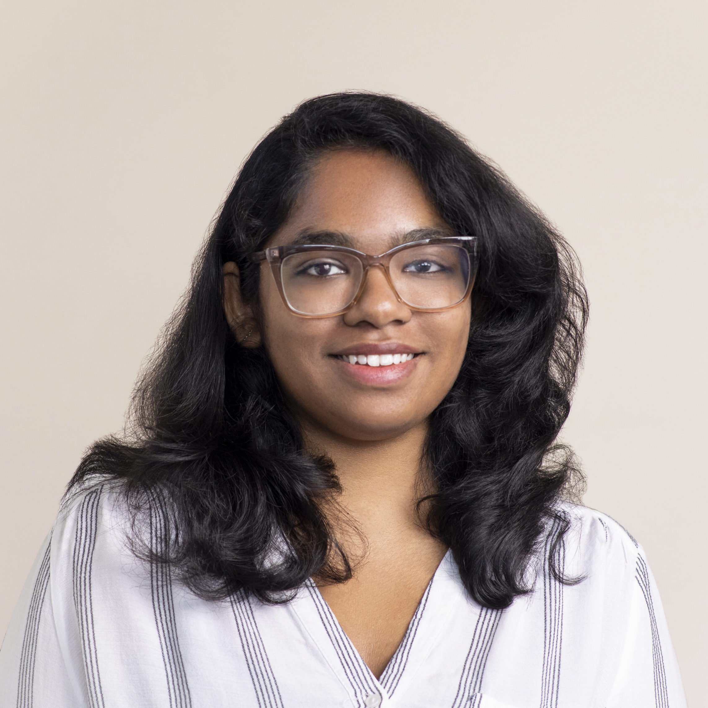 A woman with long, dark hair and glasses smiles at the camera. She is wearing a white and gray striped blouse. The background is a plain, light color.