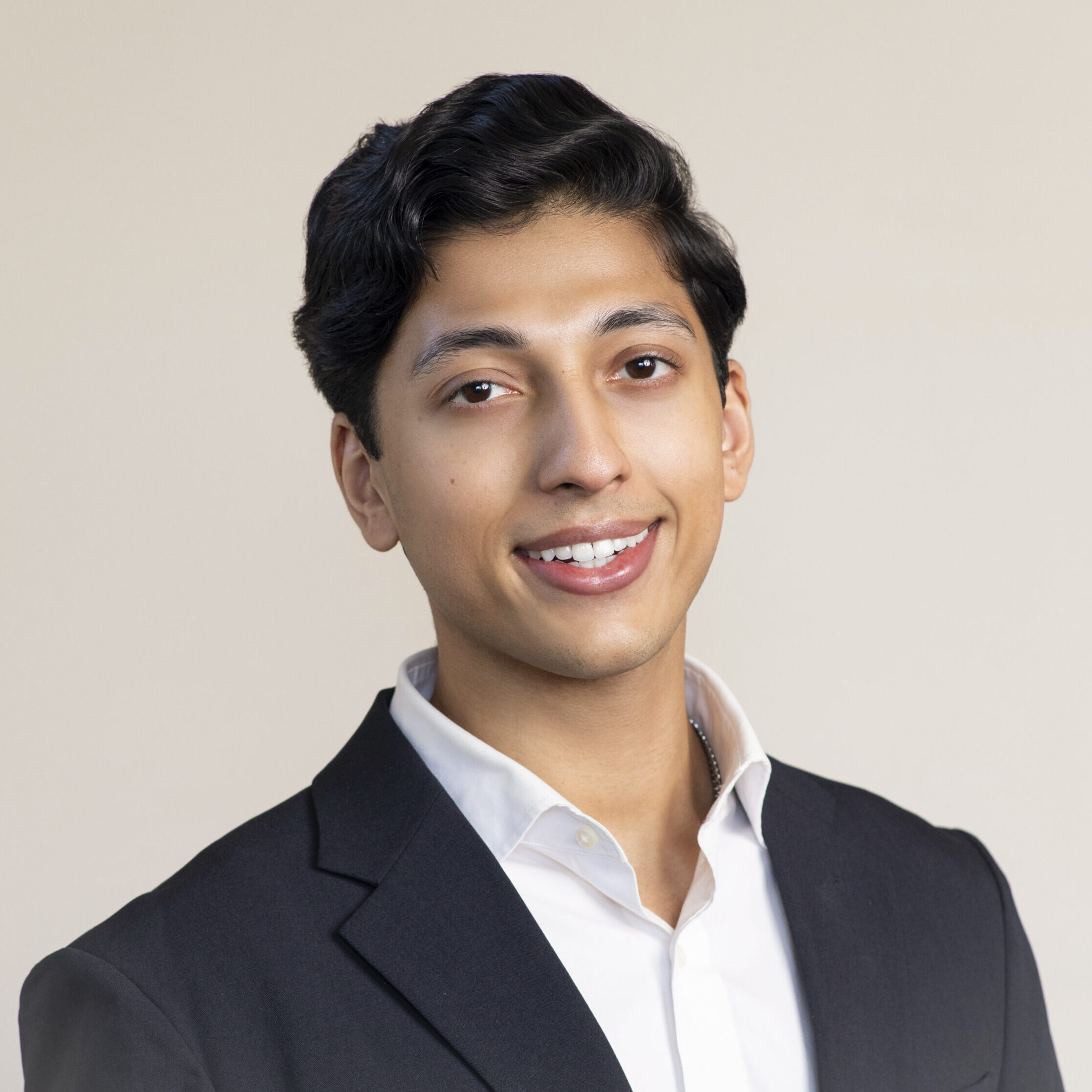 A person with short dark hair smiles at the camera. They are wearing a black suit jacket over a white collared shirt against a plain, light-colored background.