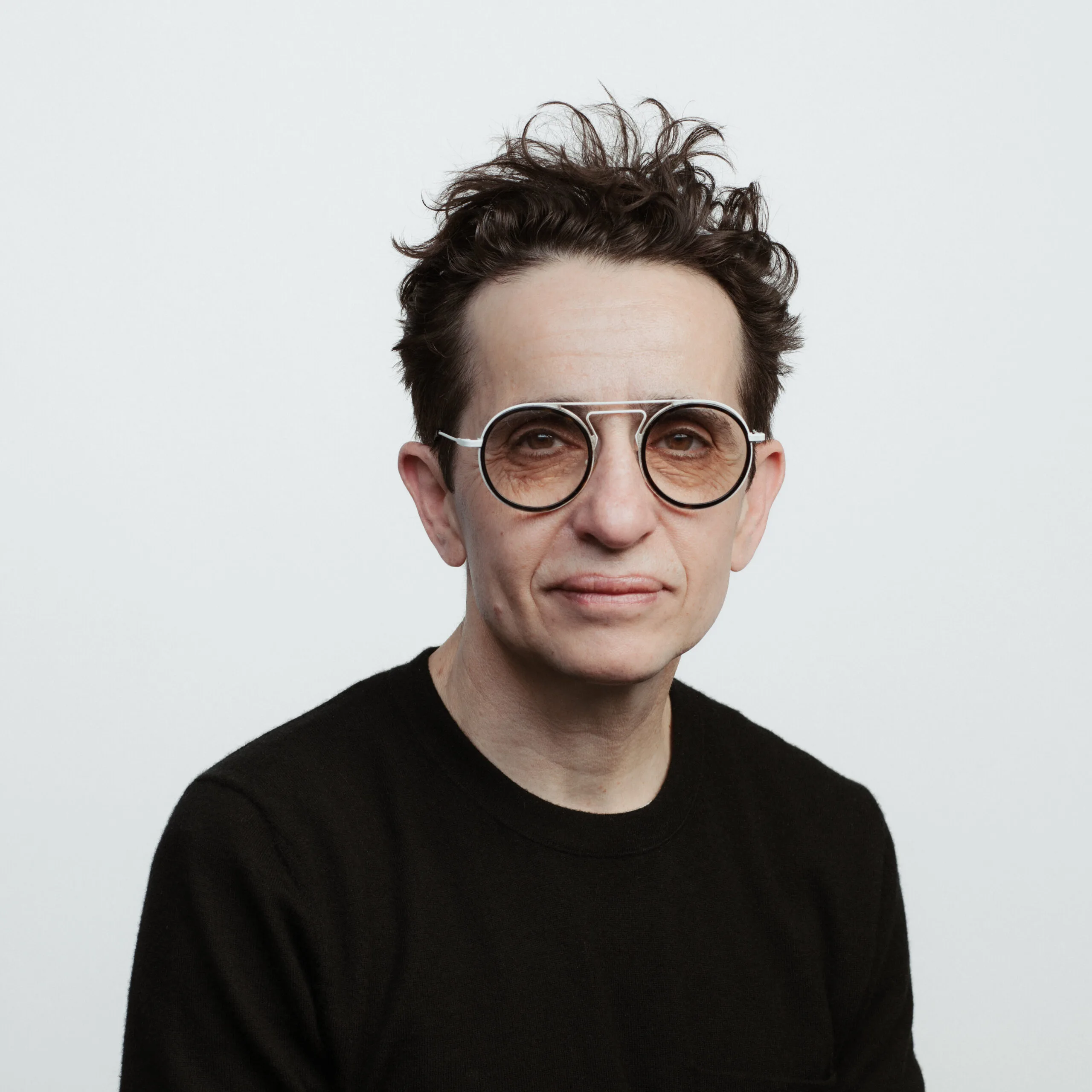 A person with short, tousled dark hair wearing large round glasses and a black sweater, poses against a plain white background, looking directly at the camera with a neutral expression.