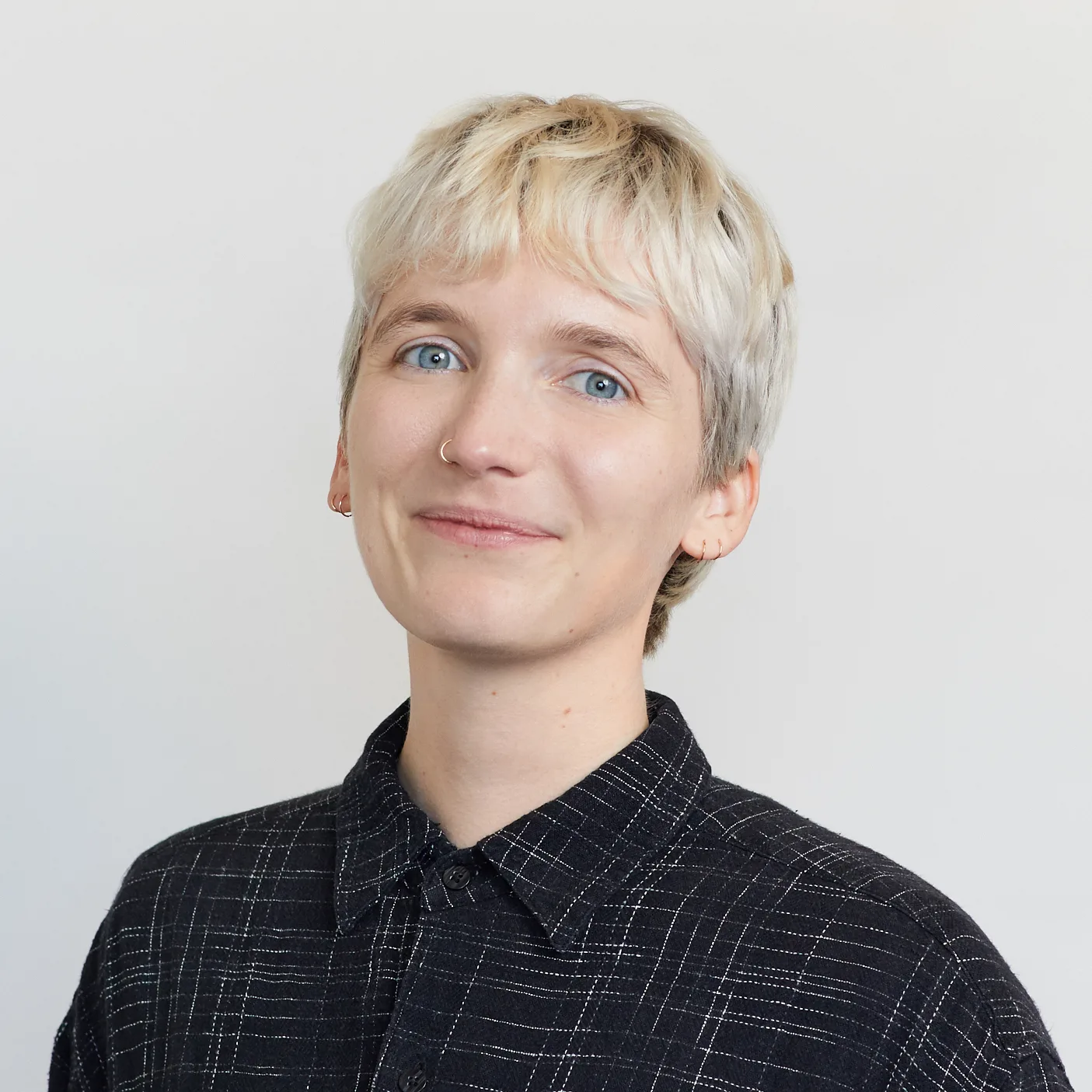 A person with short, light blonde hair and blue eyes smiles softly. They are wearing a black, checkered button-up shirt and are posed in front of a plain, light background.