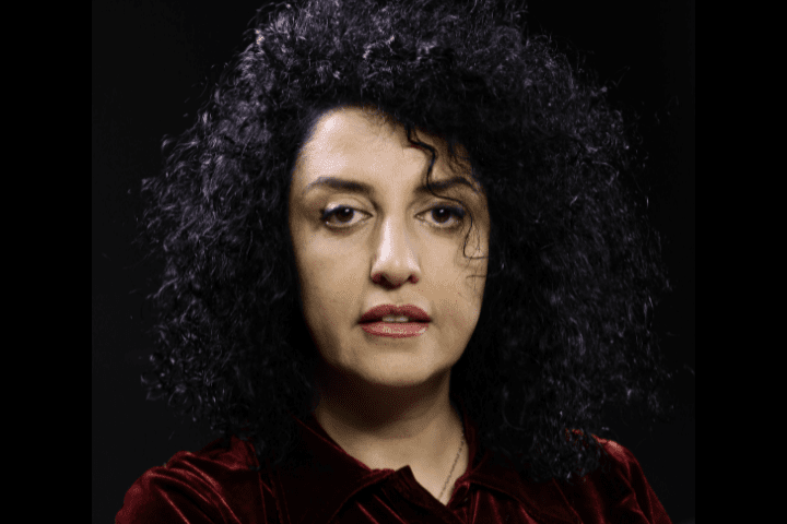 Person with curly black hair and a serious expression, wearing a dark red velvet top, against a black background.