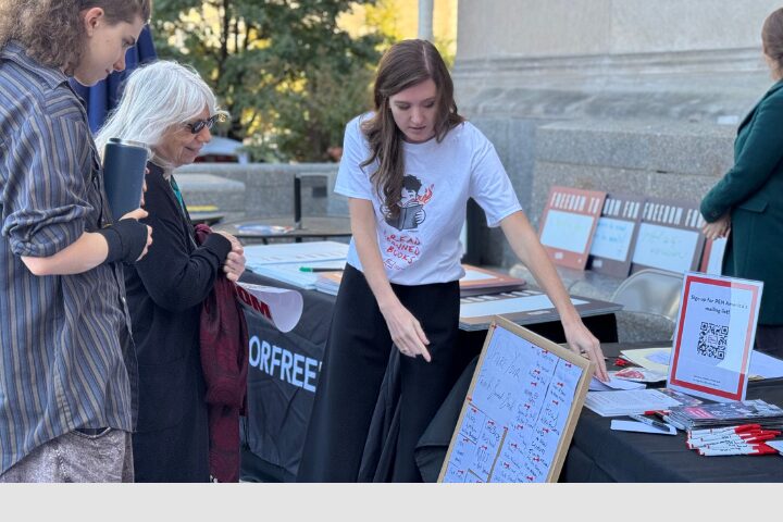 A woman in a printed t-shirt showcases a board with text and diagrams to two people at the Freedom to Read Day of Action 2024 event. A table with pamphlets and informative materials stands before her, amid the backdrop of trees and a stone building.