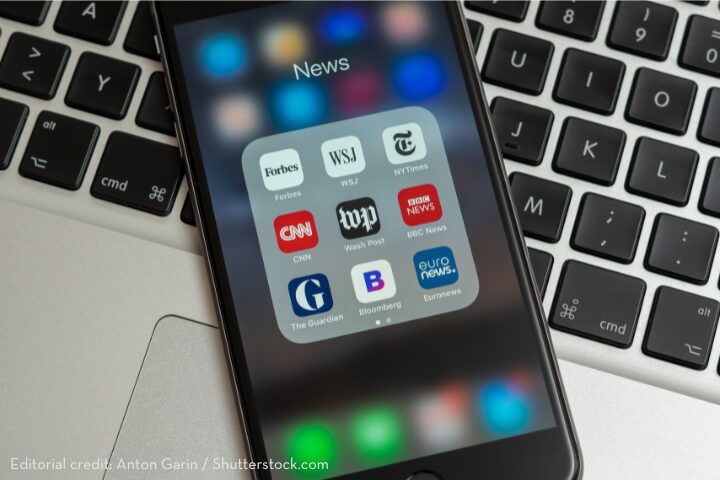 A smartphone displaying a News folder with app icons such as Forbes, CNN, The Guardian, and BBC News rests on a laptop keyboard.
