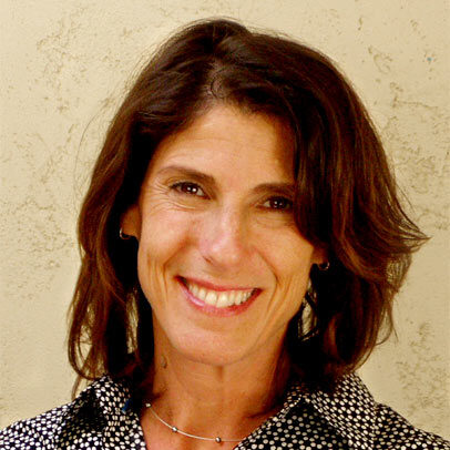 A woman with shoulder-length brown hair smiles at the camera. She is wearing a black and white polka dot blouse and a simple necklace, standing in front of a neutral, textured wall.
