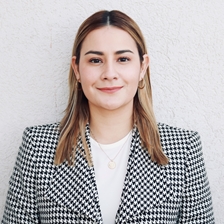 A person with shoulder-length hair wearing a black and white checkered blazer over a white top, standing against a plain white background, looking at the camera with a neutral expression.