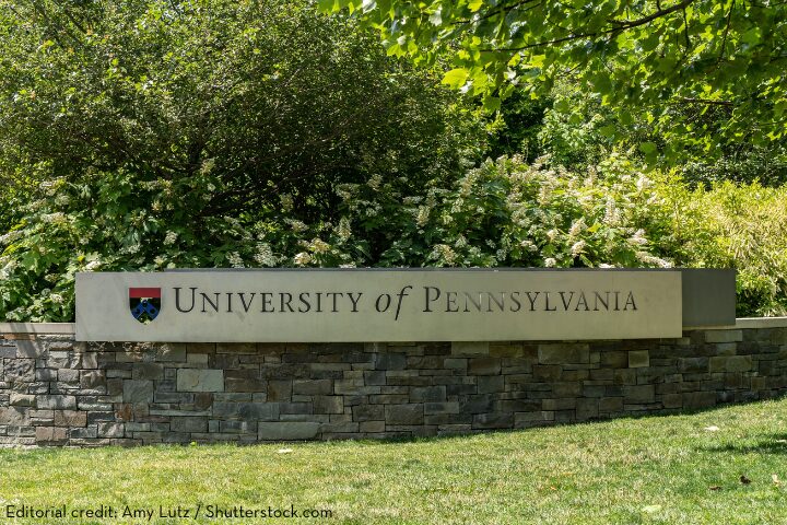 A stone sign reads University of Pennsylvania with a small emblem on the left. It is surrounded by lush green trees and plants. The grass in front is neatly trimmed.