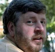 A person with short brown hair and a beard is looking over their shoulder outdoors. The background is a blurred mix of green foliage and natural light.