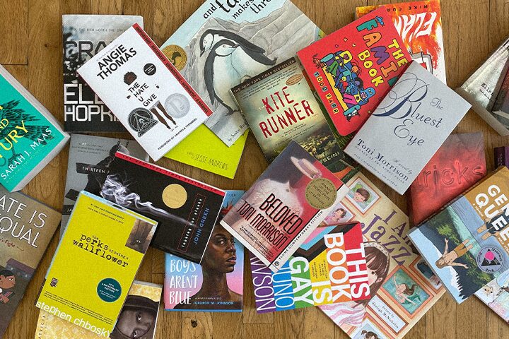 A collection of various books spread out on a wooden surface. Titles include The Bluest Eye, The Kite Runner, Beloved, and The Perks of Being a Wallflower, among others. The books cover diverse genres and themes.