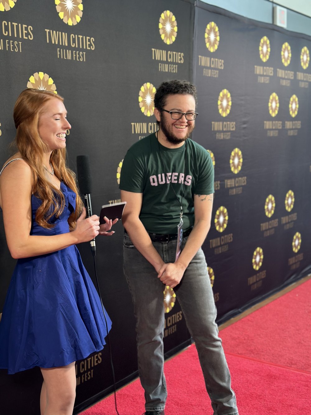 A woman in a blue dress holds a microphone, smiling as she talks to a person wearing a green Queers t-shirt and jeans. They are standing on a red carpet with a Twin Cities Film Fest backdrop.