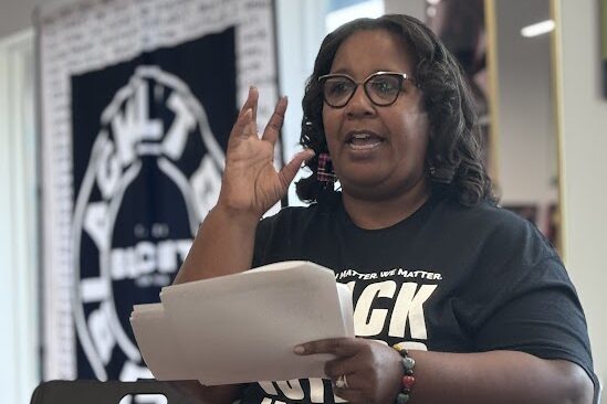 A woman with glasses, wearing a black t-shirt with white text, speaks passionately while holding papers. Behind her, a blurred banner features a circular design and text. She gestures with one hand, emphasizing her point.