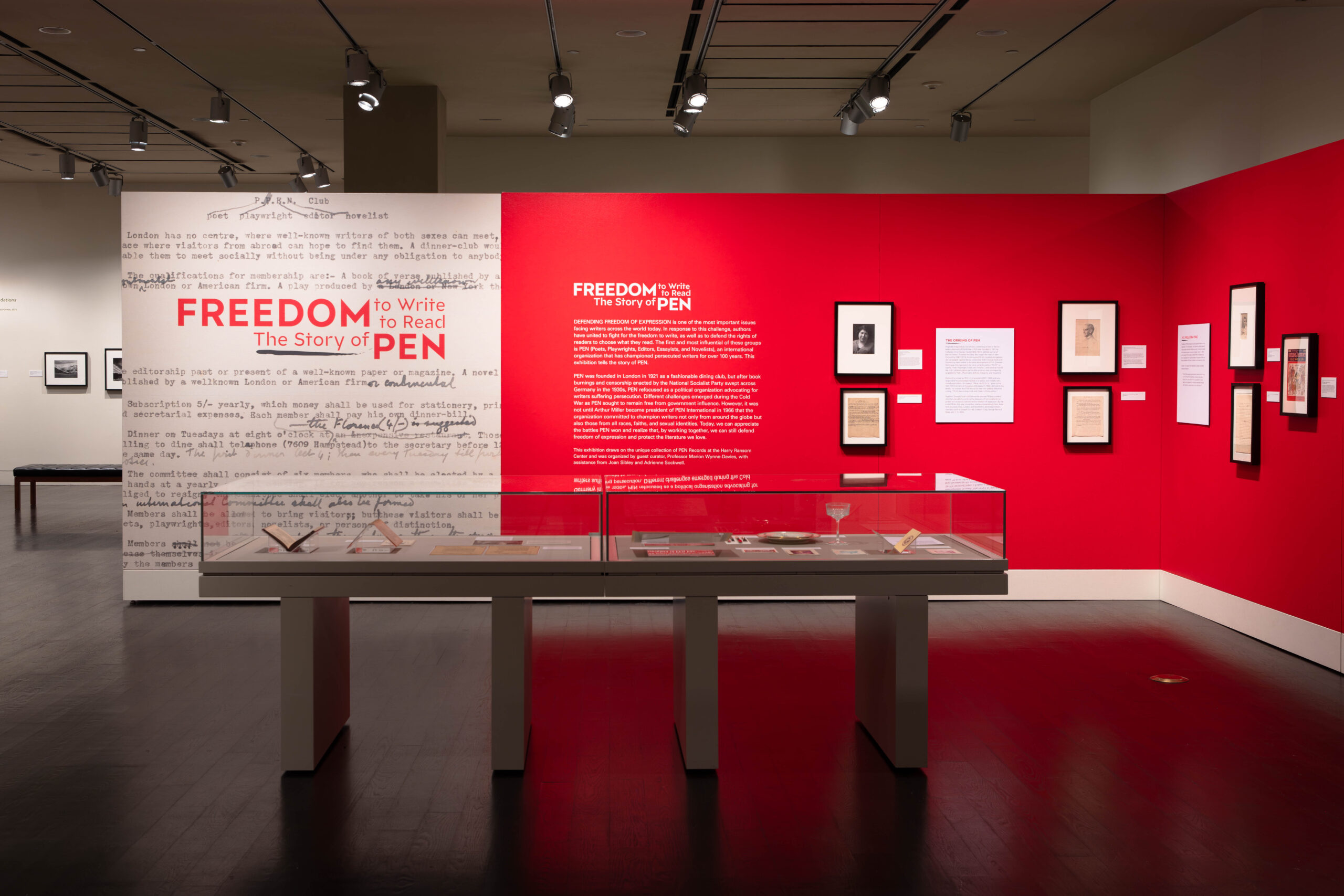 The Freedom to Write museum exhibit, set against bold red walls, artfully weaves in the story of PEN. Text panels, framed documents, and manuscripts tell its tale. A central glass display case holds priceless historical items, all illuminated by spotlights that bring each word to life.