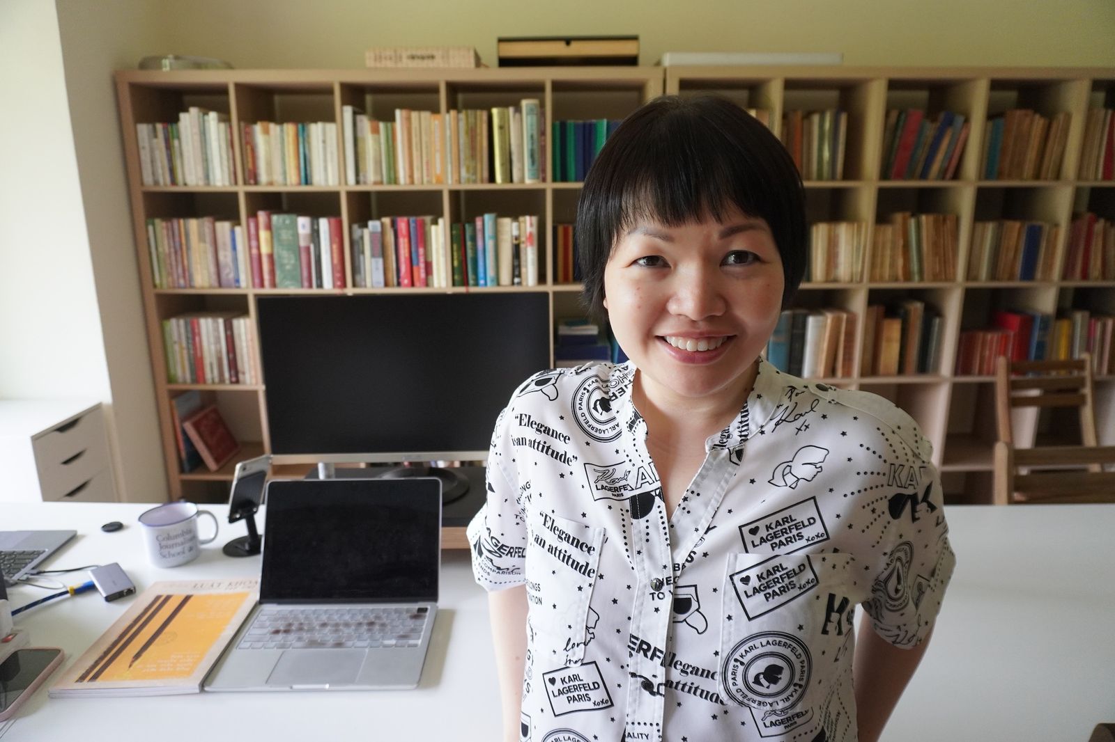 A person with short black hair smiles at the camera, standing in a room with bookshelves filled with books. On the desk in front, there are a laptop, a monitor, and some papers. The person is wearing a shirt with various patterns and text.