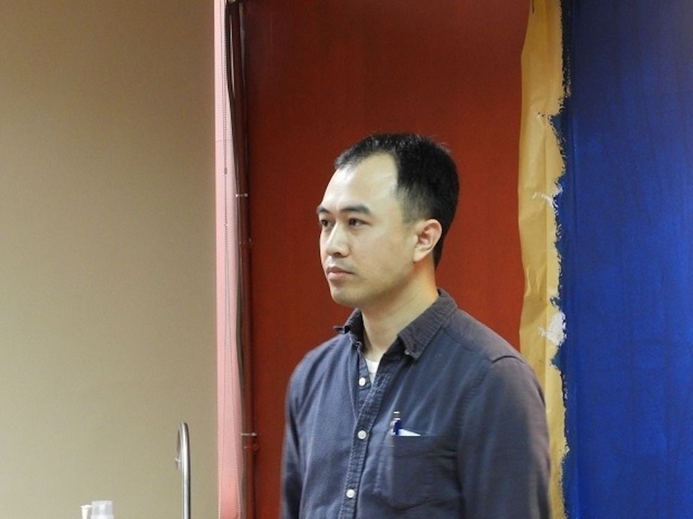 A person with short black hair stands indoors beside a colorful wall. They are wearing a dark button-up shirt and have a pen in their pocket. The background includes red, yellow, and blue vertical panels.