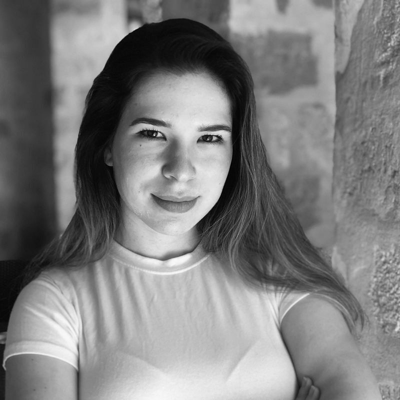 Black and white portrait of a person with long hair, wearing a light-colored shirt, standing in front of a textured stone wall. They have a neutral expression and arms crossed.