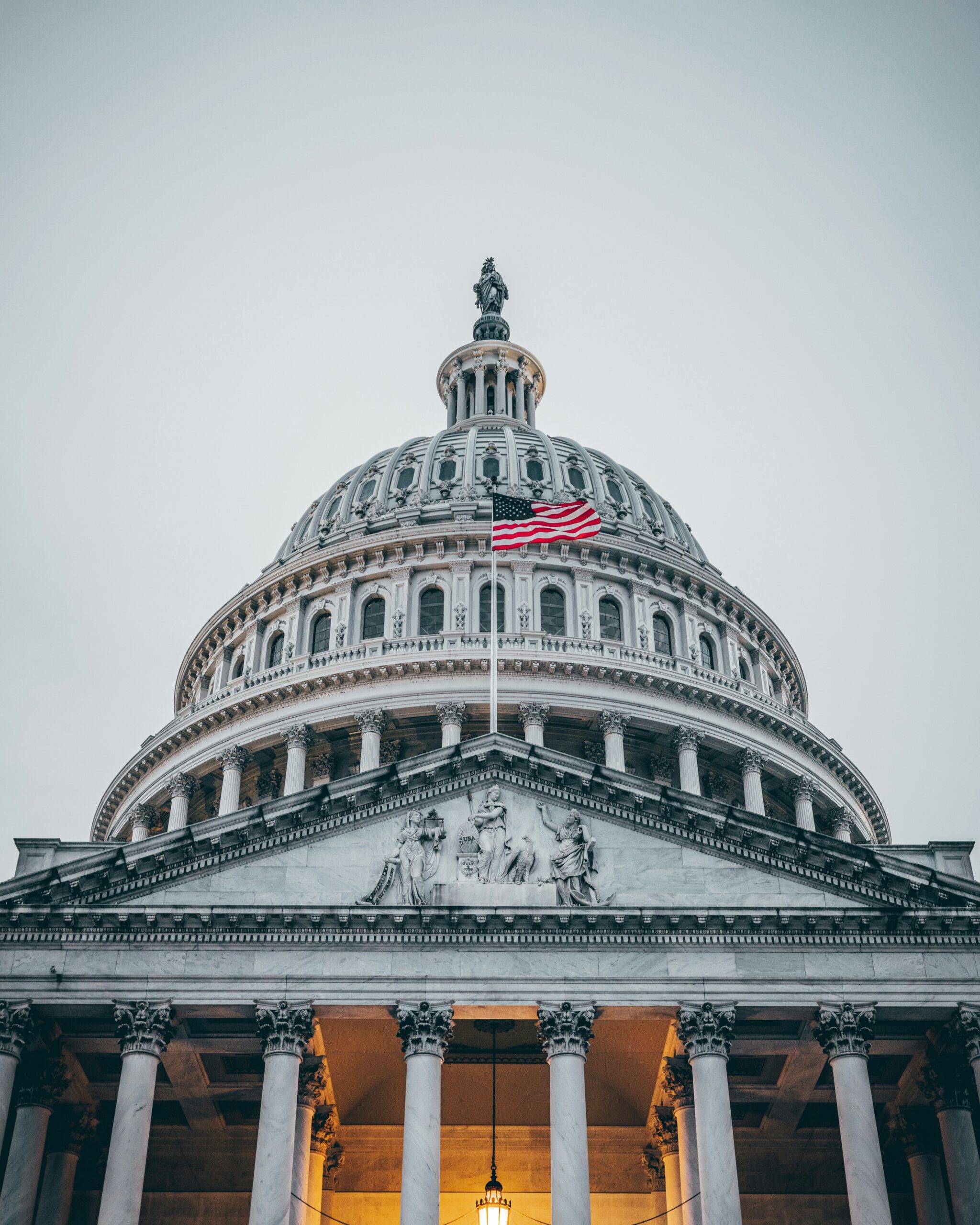 The image shows the United States Capitol Building with its iconic dome, adorned by an American flag. The buildings classical architecture features detailed columns and sculptures under an overcast sky.