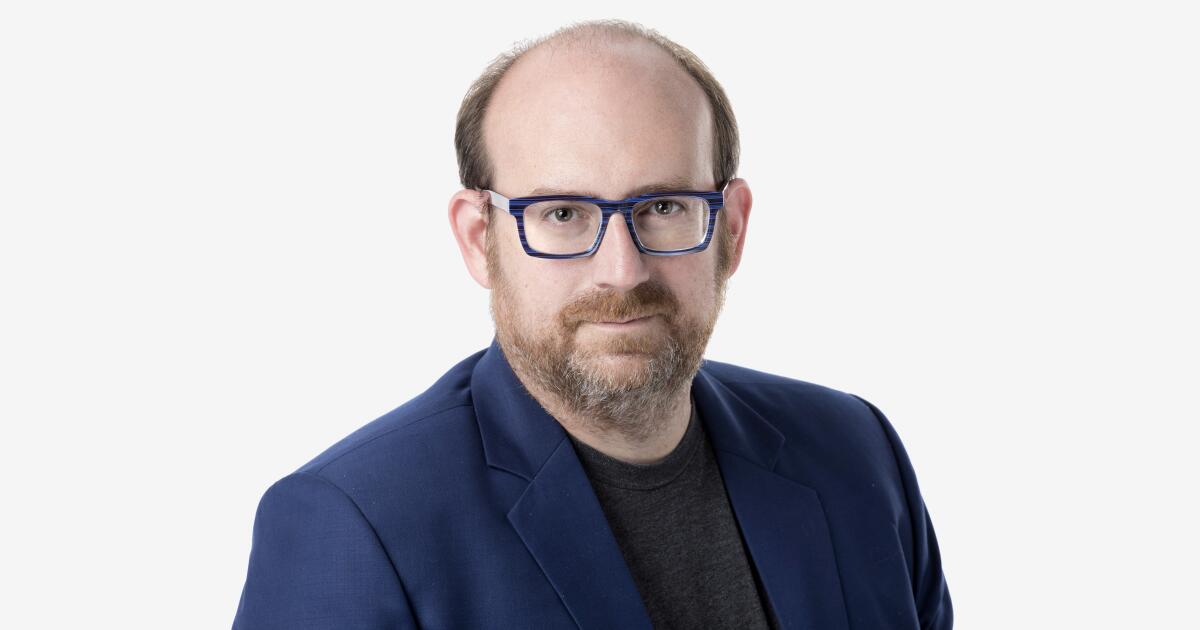 A man with glasses and a beard is wearing a blue blazer and a dark shirt. He is looking directly at the camera against a plain white background.