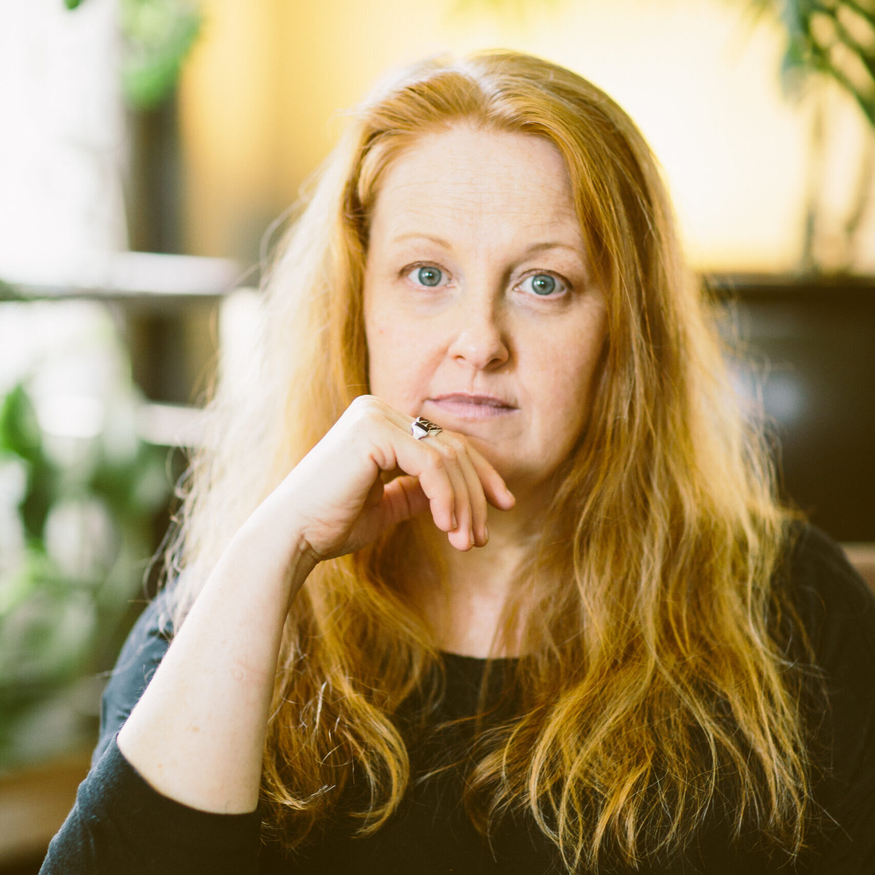 A woman with long red hair and a black shirt sits indoors with her chin resting on her hand. She has a thoughtful expression. The background is softly lit with green plants visible.