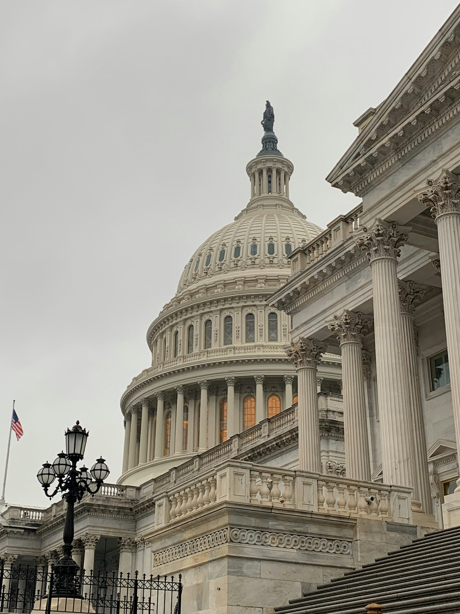 The image shows the United States Capitol building with its iconic dome and neoclassical architecture. An American flag and a decorative lamppost are visible in the foreground under a cloudy sky.