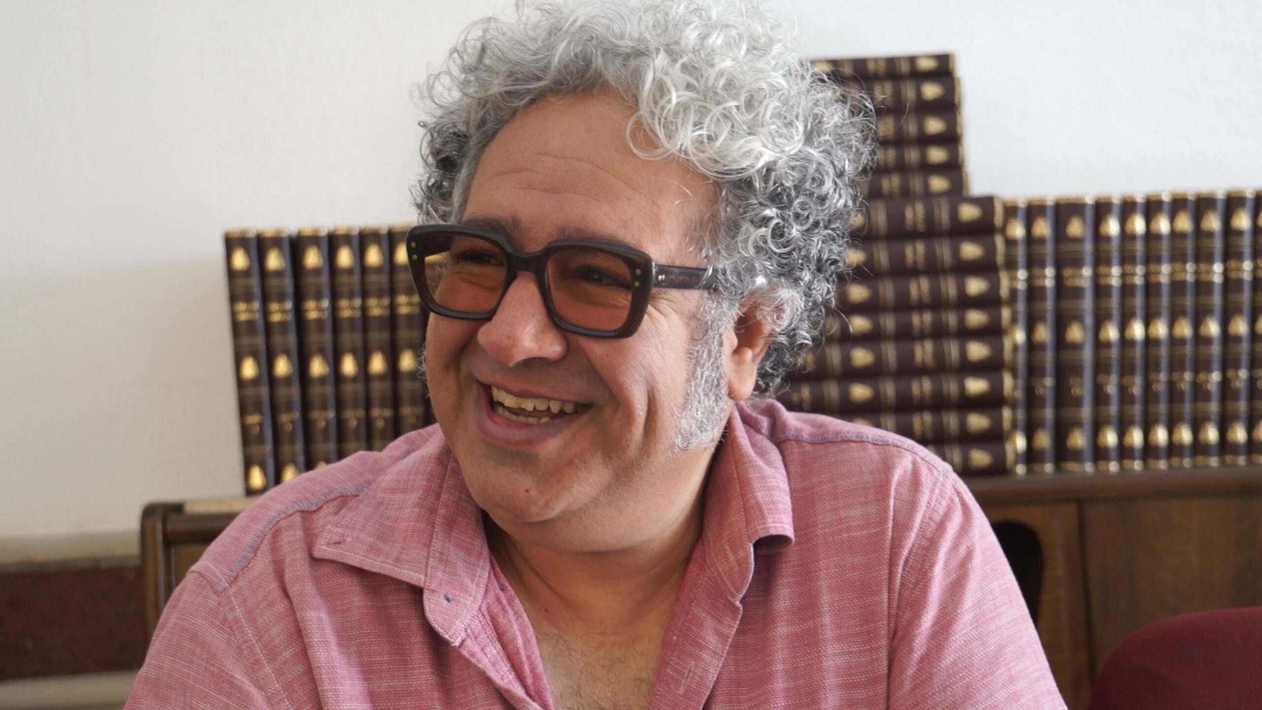 A smiling person with curly gray hair and glasses, perhaps inspired by the works of Baktash Abtin, wears a pink shirt while sitting in front of shelves filled with books.