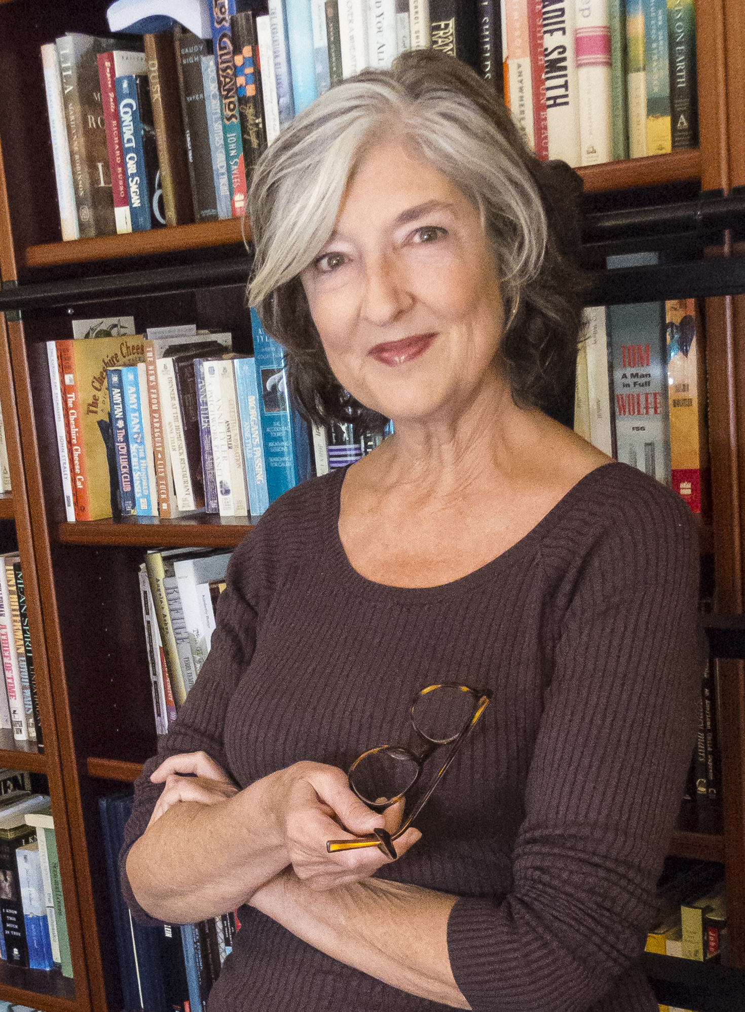 A woman with gray and brown hair smiles while standing in front of a bookshelf filled with books. She is wearing a brown top and holding glasses in her hand.