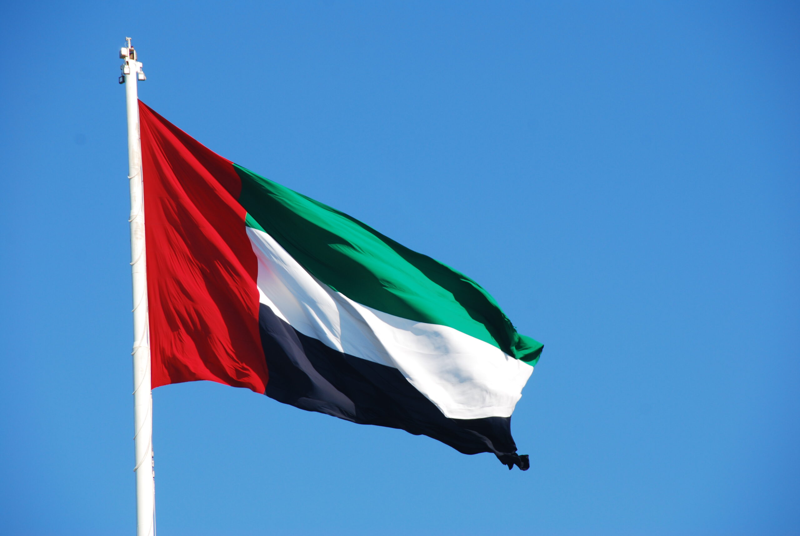 The flag of the United Arab Emirates, featuring red, green, white, and black horizontal bands, flutters against a clear blue sky.