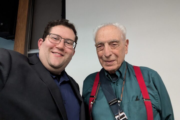 Two men pose for a photo together indoors. The man on the left wears glasses and a dark suit, while the man on the right wears a green shirt with red suspenders and a camera strap around his neck. Both are smiling at the camera.