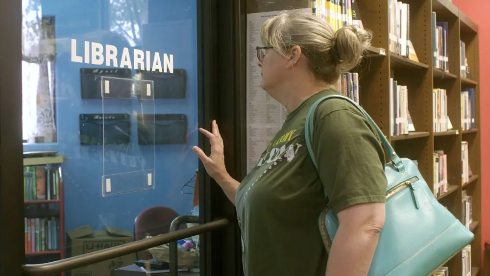 A woman with a ponytail and glasses stands outside a door labeled Librarian in a library. She is wearing a green shirt and carrying a large blue bag. Bookshelves filled with books are visible in the background.