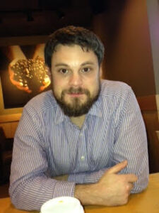 A man with a beard is sitting at a cafe table, wearing a striped shirt. Leaning slightly forward with arms crossed, he listens intently to one of the world literature speakers. A framed artwork is visible on the wall behind him.