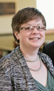 A smiling person with short hair and glasses, perhaps one of the renowned world literature speakers, wears a patterned gray sweater over a teal top and a beaded necklace. They are indoors with a blurred background.