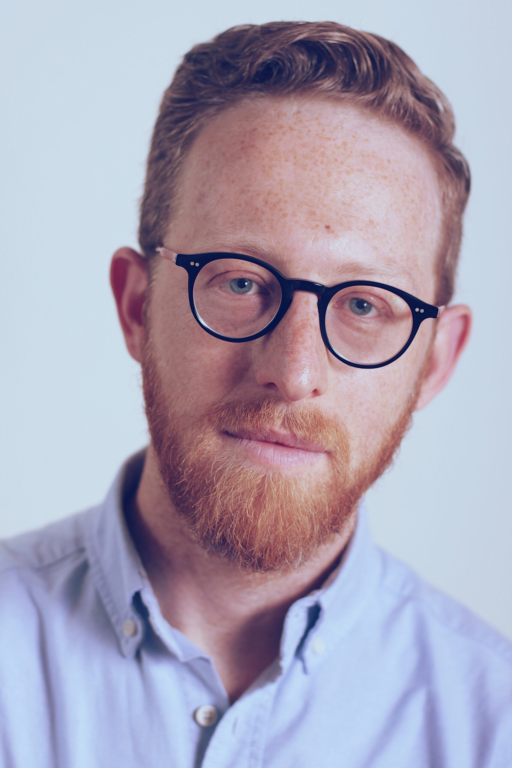 A person with short curly hair, a beard, and glasses looks directly at the camera. They are wearing a light blue collared shirt and have a calm expression. The background is plain and softly lit.
