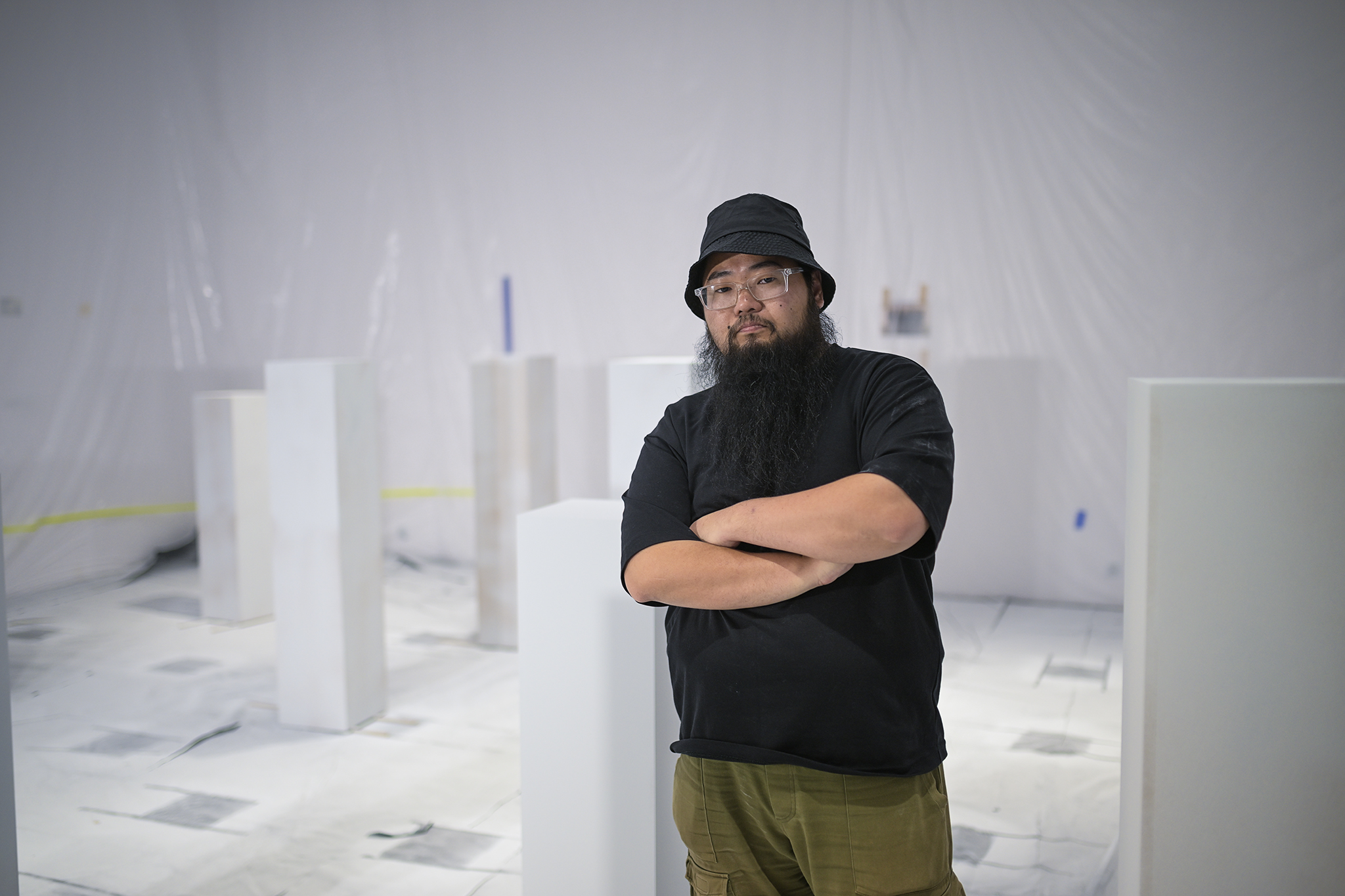 A person with a black hat and long beard stands with arms crossed in a room with white-painted columns and a plastic-covered floor and walls. The scene appears to be an art or construction space.